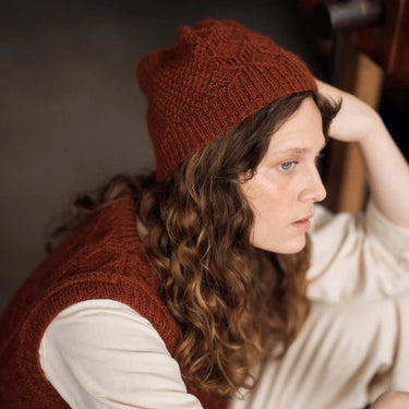 Person wearing a brown knitted hat and vest in a dimly lit room.