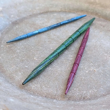 KnitPro Dreamz Wood Cable Needles with grooves to prevent slipping, shown in blue, green, and purple on a textured surface.