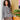 Woman wearing a stylish grey knit sweater with black beaded necklace, leaning on a dresser next to a green potted plant.