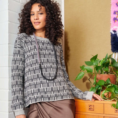 Woman wearing a stylish grey knit sweater with black beaded necklace, leaning on a dresser next to a green potted plant.