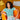 Model wearing a turquoise knit top sitting on an orange chair, with a yellow pillow and a zebra-print pillow on the sofa behind.
