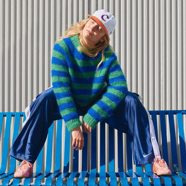 Model wearing a striped yoke sweater and trousers from Molla Mills' Hooks Out! collection, sitting on a blue bench outdoors.