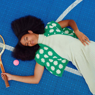 Woman in crocheted outfit from Molla Mills' Hooks Out collection, lying on a blue sports court with tennis racket and ball.