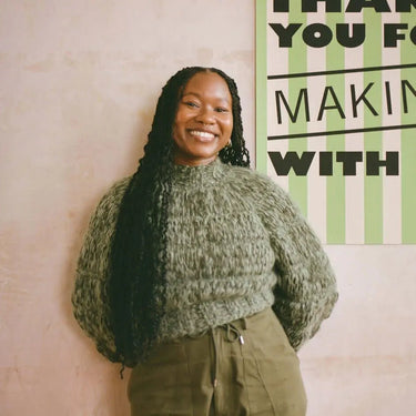 Instructor for Romanesco Bag Workshop smiling in a cozy green sweater, standing near a sign at the knitting venue.