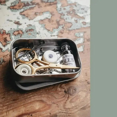 The Art of Knitwear Repair sewing kit with needles, thread, and scissors on a rustic table for garment mending.