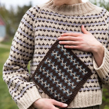 Person wearing a patterned sweater holding a knitted square outdoors. Tukubook knitting book
