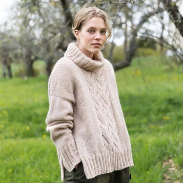 Woman wearing a beige knitted sweater in a grassy outdoor setting. Tukubook knitting book