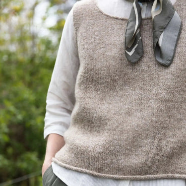 Person wearing a brown knitted vest over a white shirt with a scarf, standing outdoors. Tukubook knitting book