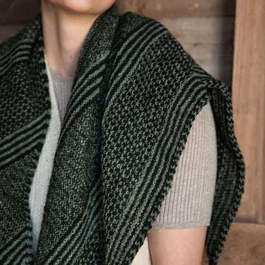 Person wearing a green knitted shawl over a beige sweater against a wooden background, Tukubook knitting book