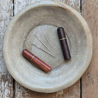 Wooden darning needles case with metal needles on rustic plate, featuring two dark brown cases, showcasing luxury and functionality.